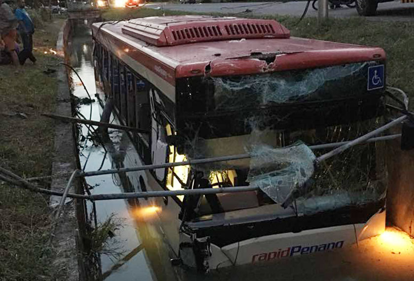 Orang ramai melihat keadaan bas yang separuh tenggelam di dalam longkang besar itu. - Foto ihsan PDRM.