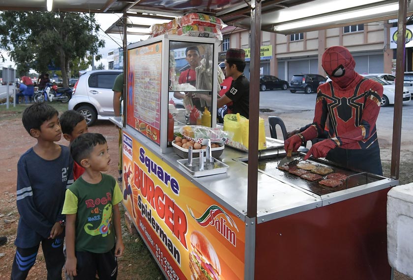 Fakhrul Fahmie Ishak, 18, (kanan) menggayakan kostum adiwira Spiderman turut membantu abangnya abangnya Ahmad Ridhuan, 22, (kiri) menyediakan pesanan burger di Kampung Raja