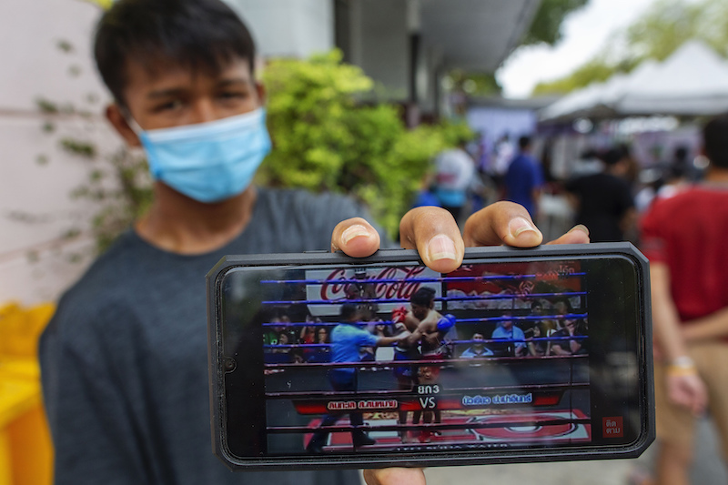 In this Thursday, March 19, 2020, photo, Rum Phonchai, a Muay Thai boxing fighter displays his mobile phone playing a video of his last fight in front of a makeshift screening facility outside Rajadamnern boxing stadium in Bangkok, Thailand. Kickboxing aficionados came from all over Thailand to attend a major Muay Thai tournament at Bangkok's indoor Lumpini Stadium on March 6, 2020. In this Thursday, March 19, 2020, photo, Rum Phonchai, a Muay Thai boxing fighter displays his mobile phone playing a video of his last fight in front of a makeshift screening facility outside Rajadamnern boxing stadium in Bangkok, Thailand. Kickboxing aficionados came from all over Thailand to attend a major Muay Thai tournament at Bangkok's indoor Lumpini Stadium on March 6, 2020. In this Thursday, March 19, 2020, photo, Rum Phonchai, a Muay Thai boxing fighter displays his mobile phone playing a video of his last fight in front of a makeshift screening facility outside Rajadamnern boxing stadium in Bangkok, Thailand. Kickboxing aficionados came from all over Thailand to attend a major Muay Thai tournament at Bangkok's indoor Lumpini Stadium on March 6, 2020.  