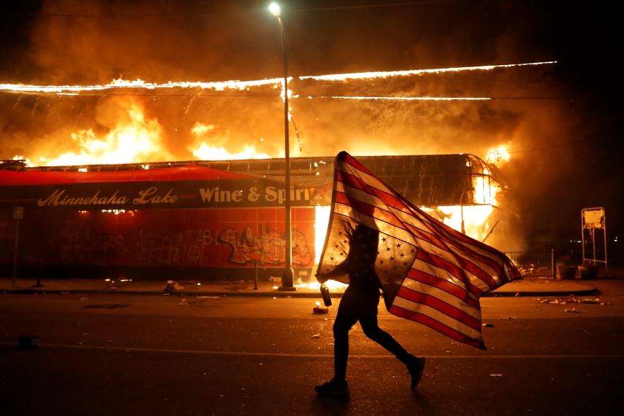 A protester carries a U.S. flag upside down, a sign of distress, next to a burning building Thursday, May 28, 2020, in Minneapolis. Protests over the death of George Floyd, a black man who died in police custody Monday, broke out in Minneapolis for a third straight night. (AP Photo/Julio Cortez)