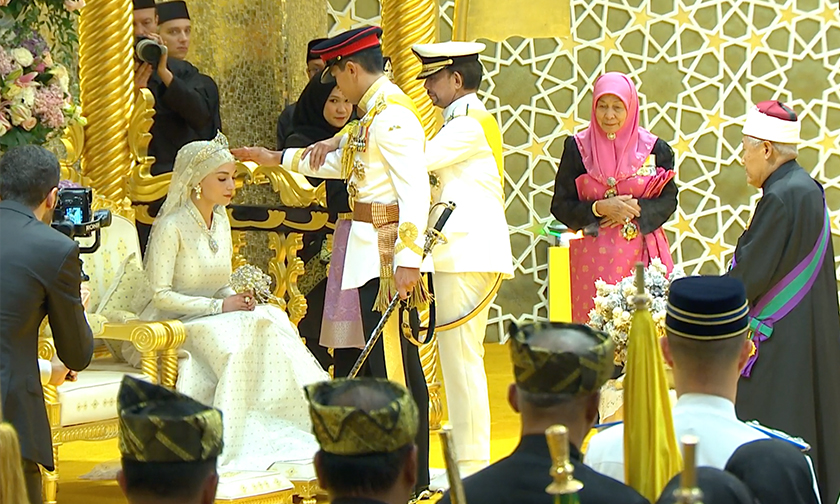 Sejurus tiba di singgahsana pelaminan, Sultan Hassanal Bolkiah berkenan memegang tangan Putera Mateen untuk diletakkan di ubun-ubun Dayang Anisha Rosnah sambil bacaan selawat kedengaran. - Putera Mateen dan Dayang Anisha selamat bersanding hari ini. - Gambar/ Tangkap layar RTBGo